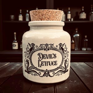 May include: A cream ceramic jar with a cork stopper. The jar has a black ornate design and the words "Devil's Lettuce". The jar is on a dark wooden surface.