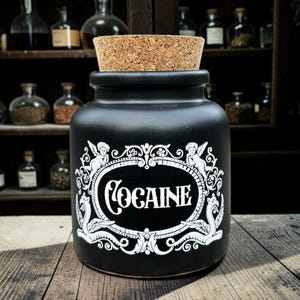 May include: Black ceramic apothecary jar with a cork stopper. The jar has a white label with the word "COCAINE" in a decorative oval frame with angel and scroll details. The jar is on a wooden surface.