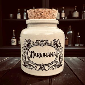 May include: A cream ceramic jar with a cork stopper. The jar has a black ornate design with the word "Marijuana". It sits on a dark wooden surface, with bottles on a shelf behind it. The jar is likely for storage.