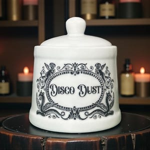May include: White ceramic jar with lid, featuring the text "Disco Dust" in a decorative oval frame. The jar is adorned with black illustrations of cherubs and ornate scrollwork. The jar is sitting on a dark wooden surface.