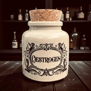 May include: A cream ceramic jar with a cork stopper. The jar has a black decorative label with the word "OESTROGEN". The label is framed by ornate scrollwork and cherub figures. The jar is on a wooden surface.