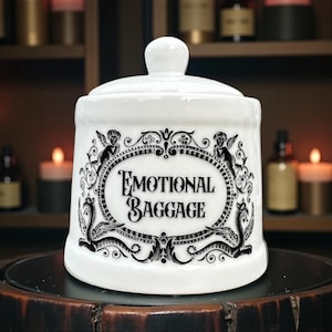 May include: A white ceramic jar with a lid, featuring the words "EMOTIONAL BAGGAGE" in a decorative oval frame. The frame includes angel and serpent illustrations. The jar sits on a dark wooden surface.