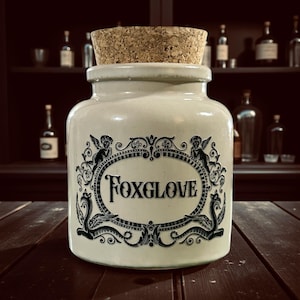 May include: A cream-coloured ceramic jar with a cork stopper, displaying the word "FOXGLOVE" within an ornate oval frame. The frame is decorated with cherubic figures and scrollwork. The jar rests on a dark wooden surface.