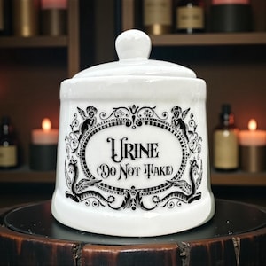 May include: White ceramic apothecary jar with lid, featuring the word "Urine" and "Do Not Take" in black script within an ornate black frame. Decorative accents include cherubs and stylized creatures.