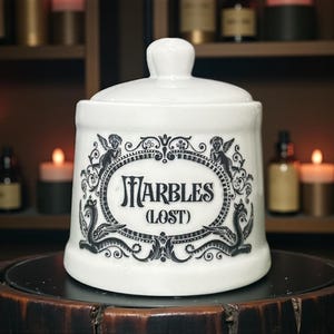 May include: White ceramic jar with a lid, decorated with a black ornate design and the words "MARBLES (LOST)". The jar is rounded and sits on a dark wooden surface. The background includes candles and bottles.