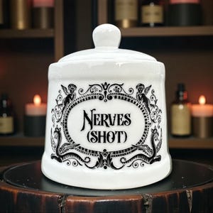May include: White ceramic apothecary jar with a lid, featuring the text "Nerves (Shot)" in a decorative oval frame with angel and serpent motifs. The jar is on a dark wooden surface.