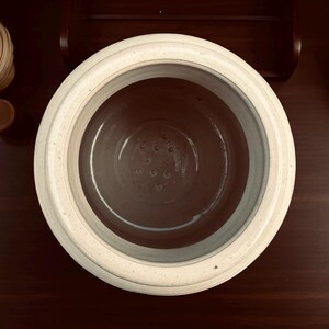May include: Overhead view of a cream-colored ceramic fermentation crock with a dark interior and small drainage holes. The crock is on a dark wood surface, with a wooden lid and other decorative wooden items in the background.