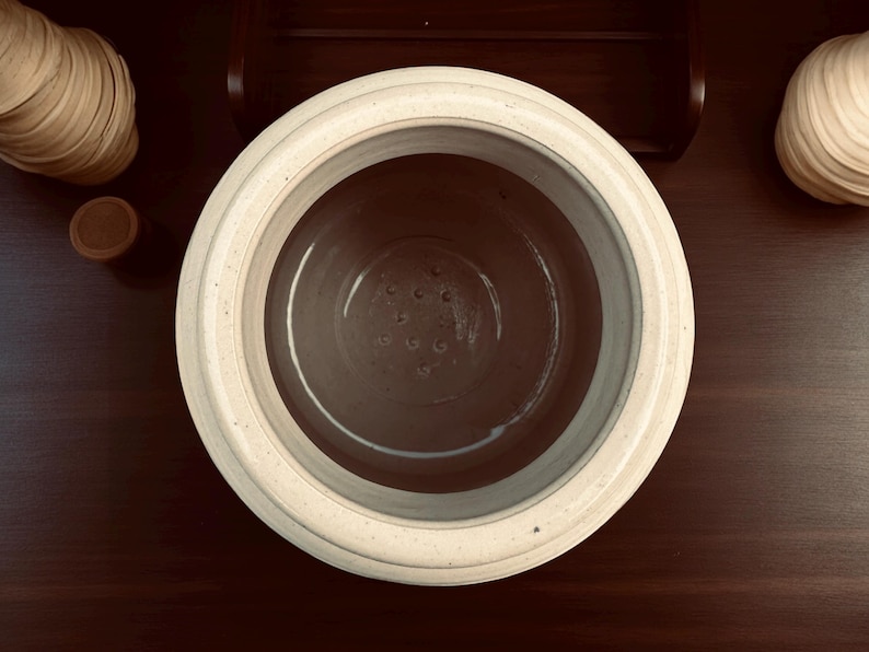May include: Overhead view of a cream-colored ceramic fermentation crock with a dark interior and small drainage holes. The crock is on a dark wood surface, with a wooden lid and other decorative wooden items in the background.