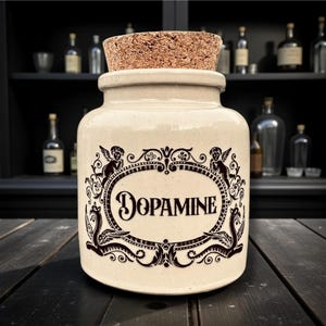 May include: A cream-coloured ceramic jar with a cork stopper. The jar has a black oval design with the word "DOPAMINE" and cherubs. It sits on a dark wooden surface. The background includes bottles on a shelf.
