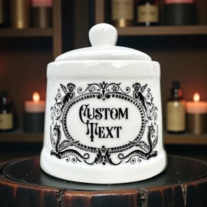 May include: White ceramic apothecary jar with a lid and a decorative black design featuring cherubs and the text "CUSTOM TEXT". The jar is displayed on a dark wooden surface.
