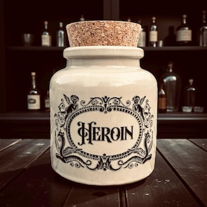 May include: A cream ceramic jar with a cork stopper. The jar features a black ornate design and the word "Heroin" in a decorative font. The jar is on a dark wooden surface.