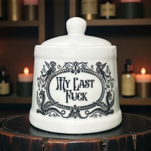 May include: White ceramic jar with a lid, featuring the text "My Last Fuck" in a decorative oval frame with angel and serpent motifs. The jar is on a dark wooden surface.