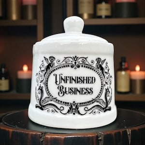 May include: White ceramic jar with a lid, featuring the text "Unfinished Business" in a decorative oval frame. The frame includes angel and serpent illustrations in black. The jar sits on a dark surface.
