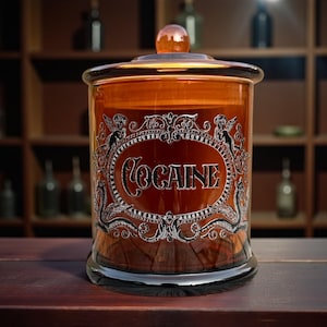 May include: An amber glass jar with a lid, displaying the word "COCAINE" in silver lettering. The jar has a decorative silver border with cherub figures. The background features shelves with bottles, creating a vintage aesthetic.