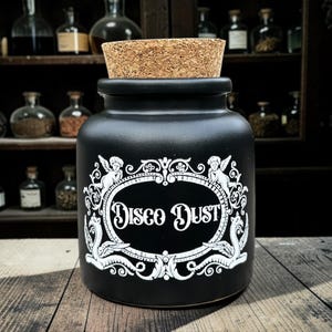 May include: A black ceramic jar with a cork stopper, labeled "Disco Dust" in a white decorative frame. The jar has a matte finish and is set against a backdrop of apothecary bottles on wooden shelves. The overall aesthetic is vintage and whimsical.