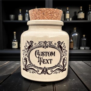 May include: A cream-coloured ceramic jar with a cork stopper. The jar features a decorative black oval with the words "CUSTOM TEXT" and ornate embellishments. The jar is on a dark wooden surface.