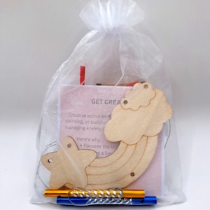 May include: A white sheer drawstring bag containing a light-coloured wooden cutout of a rainbow and star, a pink card with the text GET CREATIVE, and two small metal tubes in orange and blue. The items are visible through the translucent fabric.