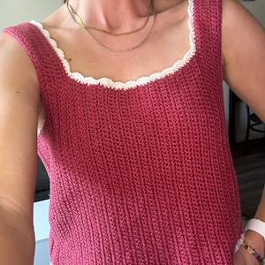 Peut inclure: Haut sans manches en crochet fait à la main, de couleur rose foncé. Le haut présente une encolure carrée et une bordure blanche festonnée le long de l'encolure et de l'ourlet. Le point de crochet crée une texture côtelée.