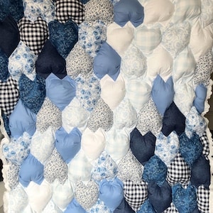 Puede incluir: Un edredón hecho a mano con un diseño en forma de corazón. El edredón presenta una variedad de telas azules, incluyendo estampados florales, cuadros vichy y mezclilla, dispuestos en un patrón texturizado. Los bordes están acabados con un ribete blanco con volantes.