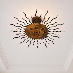 May include: An ornate ceiling light fixture in the shape of a sun. The fixture has a textured, golden-coloured central disc and curved arms radiating outwards. It is mounted on a white ceiling.