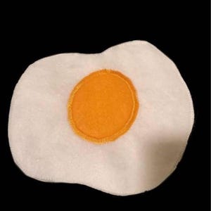 May include: A plush, white, egg-shaped item with a bright orange center, resembling a fried egg. The orange yolk is outlined with a yellow stitched border. The item is set against a black background.