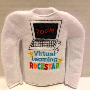 May include: White felt shirt with a laptop graphic. The laptop screen displays the word "ZOOM" in red. Below the laptop, the text "Virtual Learning ROCKSTAR" is embroidered in various colors, with yellow star accents.