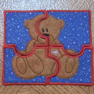 May include: A four-piece puzzle featuring a brown teddy bear design. The bear is centered on a blue background with white star-shaped accents. The puzzle pieces are outlined in red, creating a distinct border.