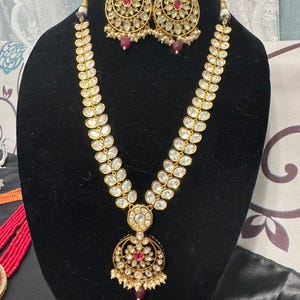 May include: A gold-toned necklace and earring set displayed on a black mannequin. The necklace features a central pendant with a red stone and a circular design. The earrings and necklace are adorned with clear stones and small pearls.