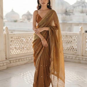 May include: A sheer bronze-toned saree featuring a delicate gold embroidered border and scattered gold embellishments. The garment is paired with a matching sleeveless blouse with thin straps, creating a cohesive and elegant ensemble.