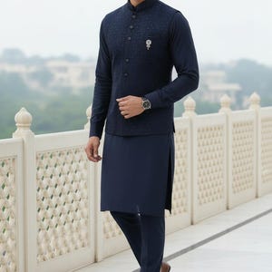 May include: Navy blue three-piece suit. The ensemble includes a long-sleeved shirt, a vest with a textured pattern and button closure, and matching pants. A silver brooch adorns the vest. The outfit is paired with a wristwatch and brown leather shoes.