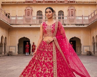 May include: A vibrant fuchsia-colored Indian lehenga with gold floral embroidery. The ensemble includes a sleeveless crop top, a flowing skirt, and a sheer dupatta. The outfit is worn in a courtyard with ornate architecture.