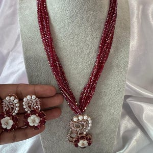 Puede incluir: Un collar de cuentas rojas de múltiples hilos con un colgante con perlas y detalles florales. También se muestran pendientes a juego con perlas y piedras rojas. El conjunto de joyas se presenta sobre una cabeza de maniquí.
