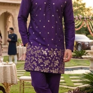 May include: A purple kurta and matching pants set. The long-sleeved kurta features gold floral embroidery at the hem and small gold dot accents. The man is wearing brown leather shoes. The outfit is suitable for a wedding or formal event.