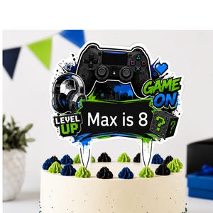 May include: A cake topper featuring a black video game controller, headphones, and a green question mark block. The design includes the text "LEVEL UP", "GAME ON", and "Max is 8" in white and green on a black banner with blue and green paint splatters.