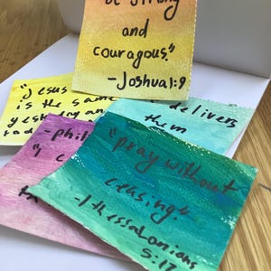 May include: A collection of colorful, hand-painted cards with inspirational quotes. The cards feature phrases like "Be Strong and courageous" and "Pray without ceasing." The color palette includes yellow, pink, green, and blue.