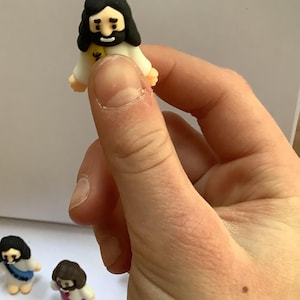 May include: A small, figurine of Jesus with black hair and a beard, wearing a white robe with a yellow emblem. The figurine is held in a hand, with other similar figurines visible in the background. The figurines are likely religious or decorative items.