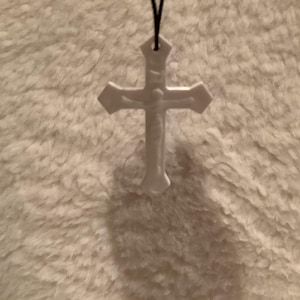 May include: A white cross pendant with a figure of Jesus Christ, suspended by a black cord. The cross has a detailed design and is set against a textured, off-white background.