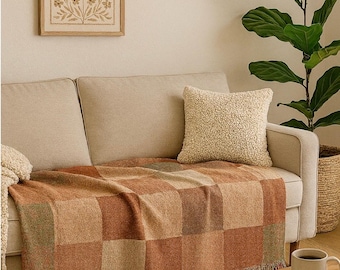 Puede incluir: Un sofá beige con una manta a cuadros en tonos marrones y verdes. Un cojín cremoso y esponjoso descansa sobre el sofá. Una obra de arte enmarcada y una planta en maceta son visibles en el fondo. Una pequeña mesa de madera con una taza y un libro.