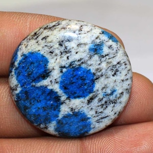 May include: A polished, round K2 stone, also known as K2 jasper, featuring a white base with black speckles and vibrant blue orb-like patterns. The gemstone is held in a hand, showcasing its natural beauty and unique color combination.
