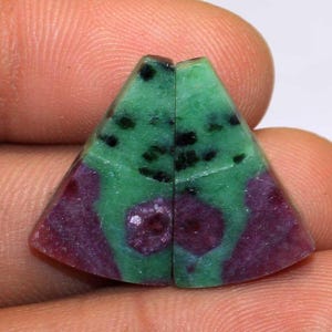 May include: Two triangular-shaped ruby zoisite gemstones. The gemstones have a vibrant green hue with black speckles and deep red-purple patches. The stones are held between fingers, showcasing their polished surfaces and natural patterns.