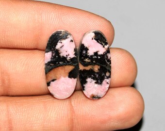 Könnte beinhalten: Zwei ovale Edelsteine mit einer Mischung aus rosa, schwarzen und braunen Farben. Die Steine werden in einer Hand gehalten, wodurch ihre polierten Oberflächen und einzigartigen Muster zur Geltung kommen. Die Edelsteine werden wahrscheinlich für Schmuck oder Dekorationszwecke verwendet.