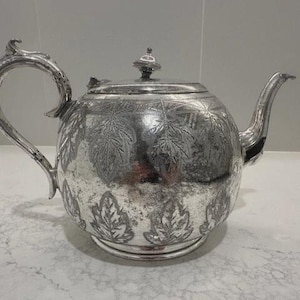 Puede incluir: Tetera plateada con diseño de hojas. La tetera tiene un cuerpo redondeado, un asa curva y un pico. La tapa tiene un pequeño pomo. La tetera está hecha de metal plateado brillante y se apoya sobre una superficie blanca.