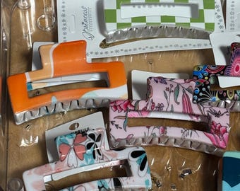 May include: A collection of colorful hair claw clips in various designs. The clips feature patterns like orange and white stripes, a green and white checkerboard, and floral prints. The clips are made of plastic and are in a clear plastic container.