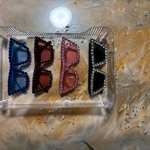 May include: Four pairs of bedazzled sunglasses in a clear plastic tray. The sunglasses are in blue, dark red, pink, and black. The frames are covered in rhinestones. The tray sits on a swirled, golden-toned surface.
