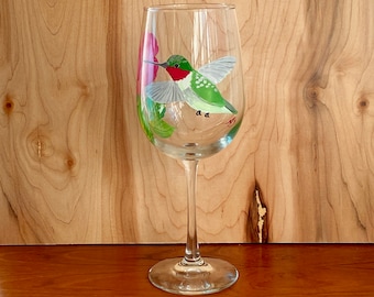 May include: A clear glass wine glass with a hand-painted hummingbird and pink flower design. The hummingbird is green, red, and white.