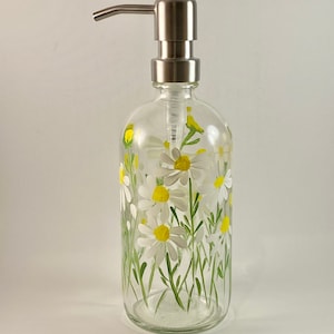 May include: A clear glass soap dispenser with a stainless steel pump. The bottle is decorated with hand-painted white daisies with yellow centers and green stems.