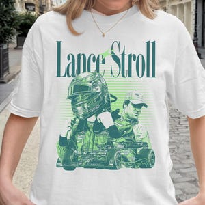 Op de afbeelding: Wit T-shirt met een groen en wit ontwerp van een autocoureur en de tekst "Lance Stroll". Het ontwerp bevat een helm, een raceauto en de naam van de coureur in een gestileerd lettertype. Het T-shirt heeft een ronde hals.