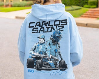 Może przedstawiać: Jasnoniebieska bluza z kapturem z nadrukiem graficznym na plecach z napisem CARLOS SAINZ, ilustracją kierowcy wyścigowego w kasku oraz bolidem Formuły 1 z numerem 55 i podpisem Carlos Sainz.