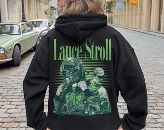 Może przedstawiać: Czarna bluza z kapturem z dużą zieloną grafiką na plecach. Wzór przedstawia napis Lance Stroll nad ilustracją kierowcy wyścigowego w kasku oraz drugim wizerunkiem kierowcy, wszystko utrzymane w odcieniach zieleni.