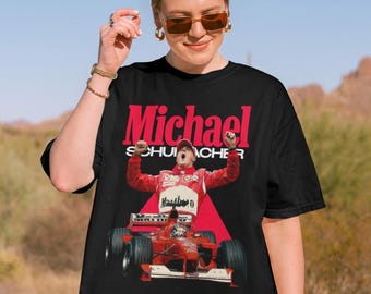 Może przedstawiać: Czarny t-shirt z krótkim rękawem z nadrukiem graficznym przedstawiającym Michaela Schumachera w czerwonym kombinezonie wyścigowym z uniesionymi w geście zwycięstwa rękami. Poniżej znajduje się czerwony bolid Formuły 1. Napis Michael Schumacher jest czerwono-biały.